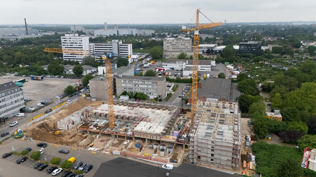 Luftaufnahme der Baustelle „Markt Carré Weingarten“ mit zwei großen Kränen, umgeben von Bürogebäuden, Straßen, einem Parkhaus und geparkten Autos an einem bewölkten Tag – das Gebiet wird derzeit erschlossen, um neuen Wohnraum zu schaffen.