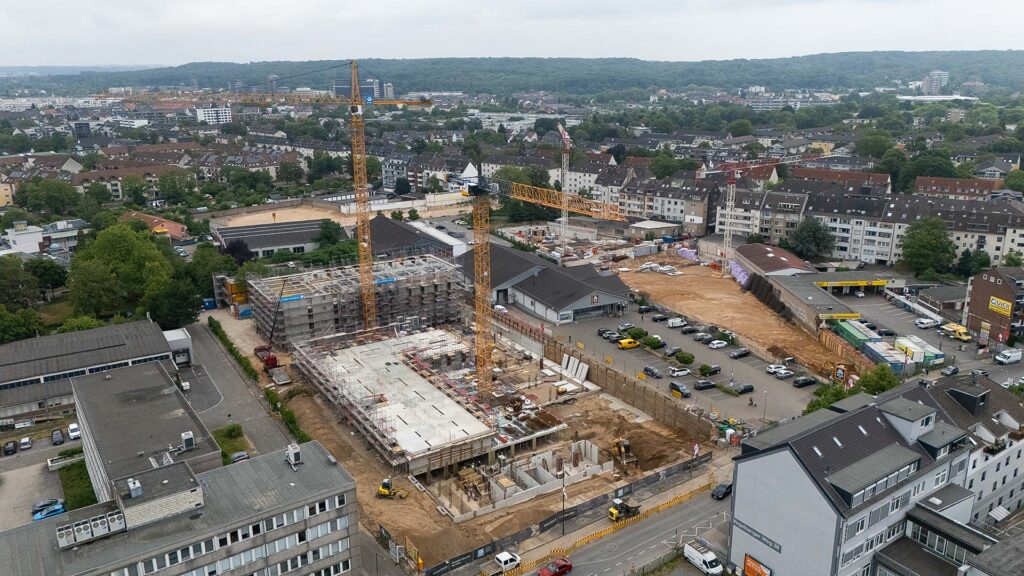 Luftaufnahme einer Baustelle am Vogelsanger Weg, auf der Kräne und teilweise fertiggestellte Bauwerke im Mittelpunkt stehen, umgeben von Straßen, Gebäuden und Bäumen unter einem bewölkten Himmel.